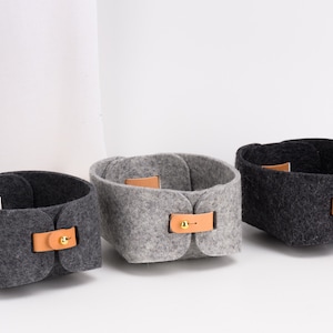 May include: Three felt storage baskets with leather handles and brass accents. The baskets are in gray, light gray, and black.