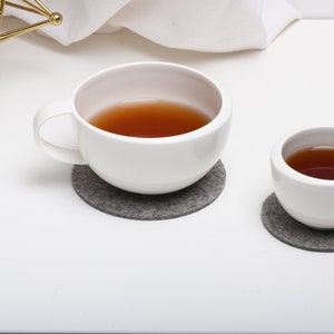 May include: Two white ceramic mugs with brown tea inside, sitting on gray felt coasters. The mugs are on a white surface.