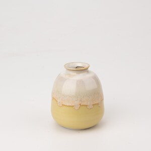May include: A small, round ceramic vase with a white and yellow glaze. The vase has a slightly rounded bottom and a wide opening at the top.