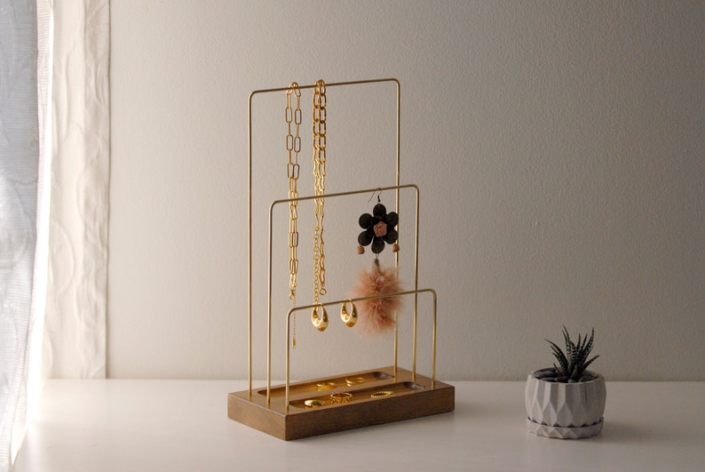 May include: A gold-coloured jewellery display stand with three tiers, holding necklaces, earrings, and rings. The stand is made of metal and wood, with a small succulent plant in a geometric pot to the right.