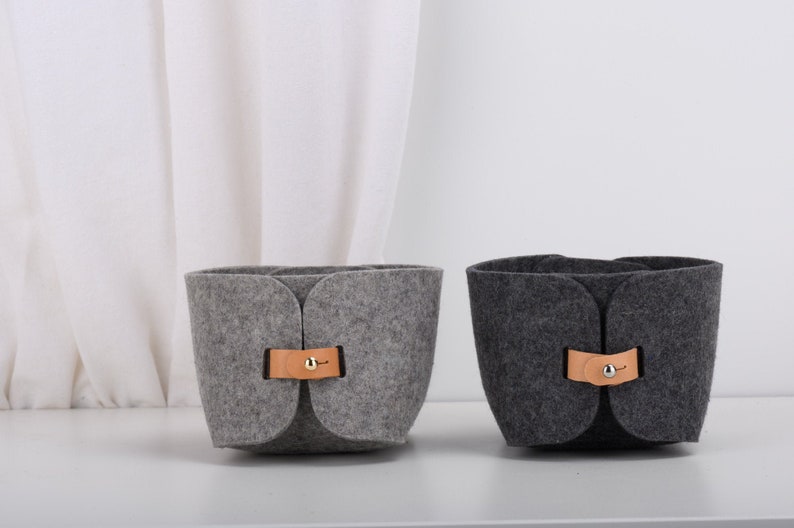 May include: Two felt storage baskets with leather handles and metal accents. The basket on the left is gray and the basket on the right is dark gray.
