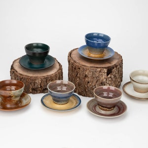 Può includere: Sei ciotole in ceramica con piattini abbinati in varie tonalità di blu, marrone e verde. Le ciotole sono esposte su ceppi di legno.