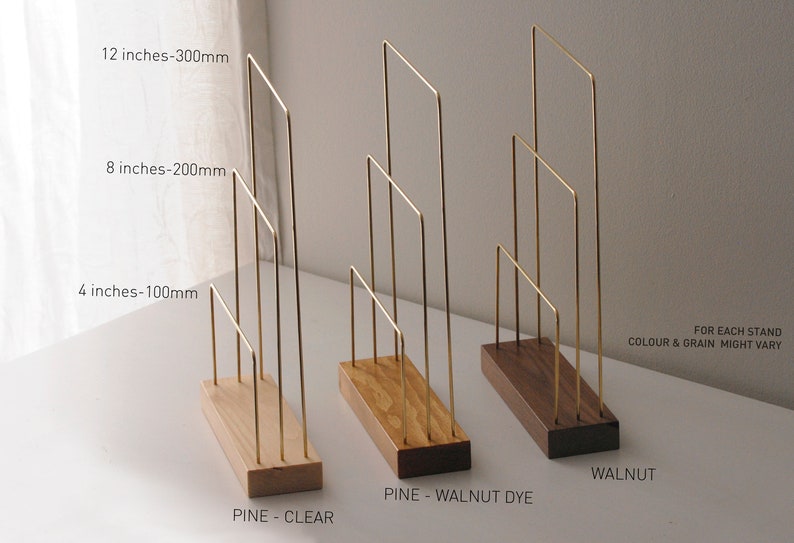 K&ouml;nnte beinhalten: Drei Pr&auml;sentationsst&auml;nder aus Messingdraht mit Holzsockeln. Die St&auml;nder sind 10 cm, 20 cm und 30 cm hoch. Die Sockel sind aus Kiefernholz in klaren und Nussbaum-Farbt&ouml;nen sowie aus Nussbaumholz gefertigt. Der Text "FOR EACH STAND COLOUR & GRAIN MIGHT VARY" ist auf dem Bild sichtbar.