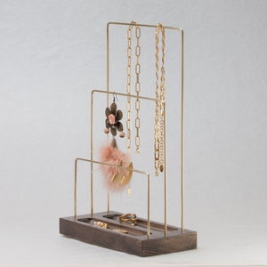 May include: A gold-coloured metal jewellery display stand with a dark wood base. The stand has multiple levels for hanging necklaces and earrings, and a tray for rings. Various gold necklaces, earrings, and rings are displayed on the stand.