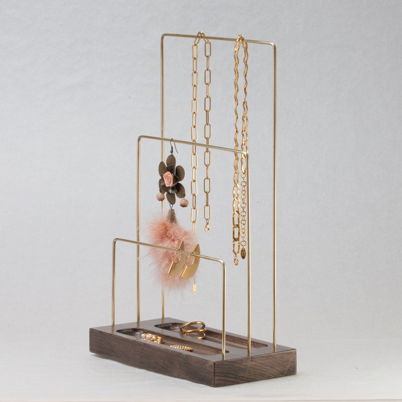 May include: A gold-coloured metal jewellery display stand with a dark wood base. The stand has multiple levels for hanging necklaces and earrings, and a tray for rings. Various gold necklaces, earrings, and rings are displayed on the stand.