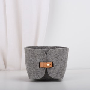 May include: A gray felt storage basket with a brown leather handle and a gold metal buckle. The basket is sitting on a white surface.