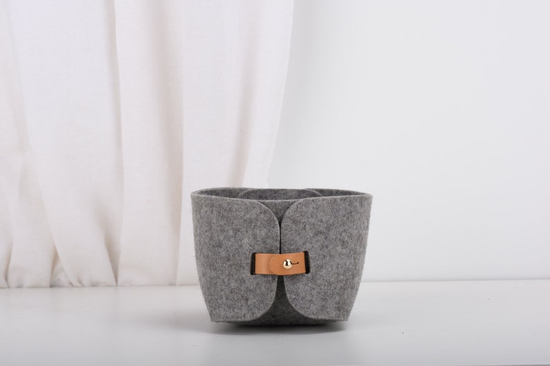 May include: A gray felt storage basket with a brown leather handle and a gold metal buckle. The basket is sitting on a white surface.