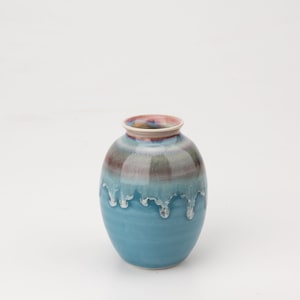 Könnte beinhalten: Eine kleine, runde Keramikvase mit blauer Glasur und einem rosa-weißen Streifen am oberen Rand. Die Vase hat einen Tropf-Glazureffekt.