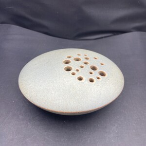 Peut inclure: Un crapaud de fleur rond en céramique grise avec de multiples trous. L'outil d'arrangement floral a une forme plate en forme de disque avec un bord légèrement surélevé. Les trous sont de tailles variables et sont disposés en motif.