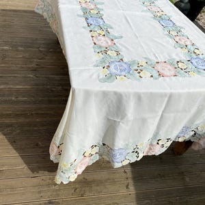May include: White tablecloth with a floral embroidered border. The border features pink, blue, and yellow flowers with green leaves.