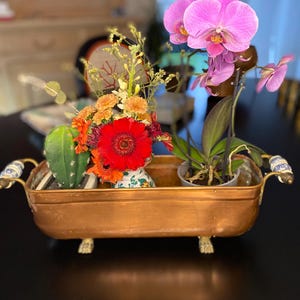 May include: A copper-colored metal planter with floral arrangements and a potted orchid. The planter has decorative handles and feet. The floral arrangement includes a red gerbera daisy, orange flowers, and greenery. The orchid has vibrant pink blooms.