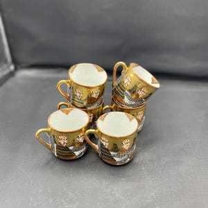 Op de afbeelding: Set van zes gouden gedetailleerde porseleinen theekopjes met een zwart en gouden ontwerp met een herhaald beeld van een mannengezicht.