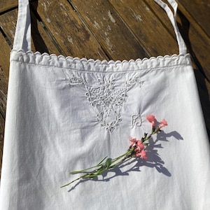 May include: White apron with embroidered floral details and scalloped trim. The apron has long straps and is adorned with a sprig of pink flowers. The apron is made of a soft, lightweight fabric.