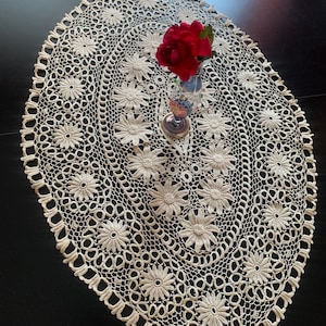 Puede incluir: Un tapete de ganchillo de color crema con forma ovalada que presenta un patrón floral repetitivo con motivos de margaritas y bordes de encaje calado. Un pequeño jarrón de cristal con una rosa artificial de color rojo intenso.