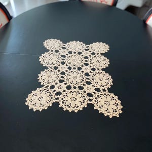 Peut inclure: Nappe en crochet beige avec un motif floral sur une table ovale noire. La nappe est faite d'un tissu délicat à tissage ouvert et présente une série de motifs floraux interconnectés. La table est entourée de chaises avec des motifs corail.