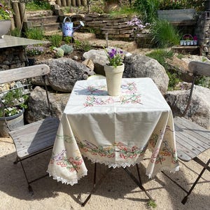 May include: A small square table with a cream-colored tablecloth featuring embroidered birds and floral designs. The table is set with a small flower pot and is flanked by two wooden chairs. The scene is set outdoors in a garden.