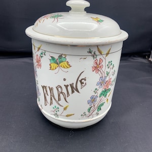 Peut inclure: Un pot en céramique blanc avec un couvercle, orné de motifs floraux et de papillons colorés. Le mot "FARINE" est écrit en script brun. Le pot a une bordure dorée.