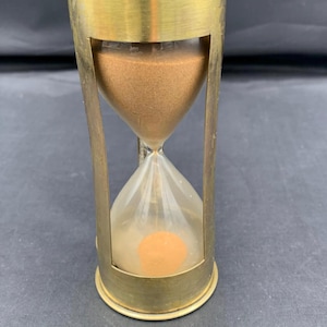 Op de afbeelding: Een zandloper met een cilindrisch messing frame met een metallic afwerking. De glazen bol bevat fijn, lichtbruin zand, waarbij een deel van het zand zich in de onderste kamer bevindt en de rest in de bovenste kamer.