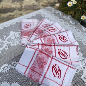 Op de afbeelding: Een stapel witte linnen servetten met rode bloemen- en ruitpatronen. Elke servet heeft een rood monogram. De servetten zijn gevouwen en gerangschikt op een wit kanten tafelkleed.