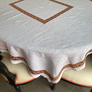 Peut inclure: Nappe ovale blanche avec une bordure brodée géométrique marron et blanche. La nappe a un motif brodé carré au centre.