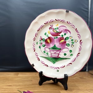 Könnte beinhalten: Dekorative Keramikplatte mit gewelltem Rand, mit handgemaltem Design eines Hahns, Blumen und einer dekorativen Vase in Rosa-, Grün- und Lilatönen. Die Platte wird auf einem schwarzen Ständer präsentiert.