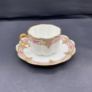 Peut inclure: Un ensemble tasse et soucoupe blanc avec un bord festonné. La tasse et la soucoupe sont décorées de motifs floraux roses et de bordures dorées. La tasse a une anse dorée. L'ensemble est sur fond sombre.