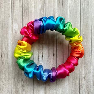 May include: A rainbow-colored scrunchie made of satin fabric. The scrunchie is in the shape of a circle and is lying flat on a wooden surface.