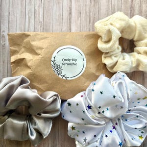 May include: An assortment of scrunchies in different materials and shades. Features a silver satin scrunchie, a white scrunchie adorned with colourful stars, and a cream-coloured terry cloth scrunchie. A brown paper package with a "Lucky Dip Scrunchie" sticker is also visible.