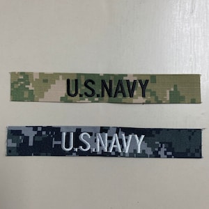 May include: Two fabric patches with a camouflage pattern. The top patch is tan and green with black text that reads "U.S. NAVY". The bottom patch is gray and blue with white text that reads "U.S. NAVY".