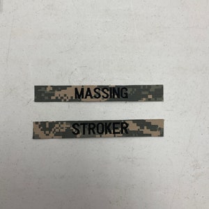 May include: Two camouflage fabric name tags with black text. The top tag reads "MASSING" and the bottom tag reads "STROKER".