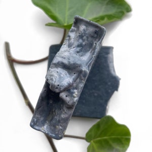 May include: Two bars of charcoal soap, one with a textured surface and the other with a smooth surface. The soap is a dark gray color with white flecks.