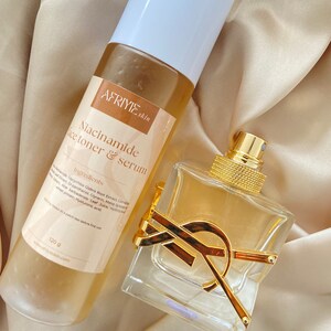 Puede incluir: Una botella de perfume de vidrio transparente con un logotipo YSL dorado. La botella está sobre una superficie de tela marrón claro. Junto a la botella hay una botella de plástico transparente con una tapa blanca. La botella está etiquetada con "AFRIYIE skin Niacinamide face toner & serum" e "Ingredientes".