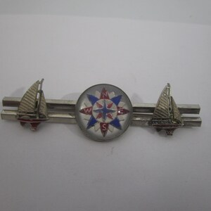 May include: A silver-toned tie bar featuring a central circular compass rose emblem with blue, red, and white enamel details. The compass is flanked by two small silver-toned sailboat figures, each with a mast and sail, positioned on the horizontal bar.