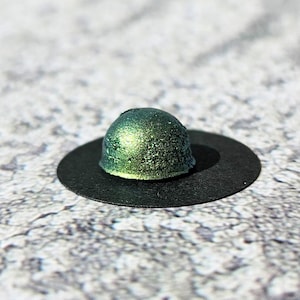 May include: A small, dome-shaped object with a shimmering, metallic green finish, centred on a flat, circular black base. The surface of the dome has a textured, glittery appearance that reflects light, creating a distinct contrast against the matte black disc.