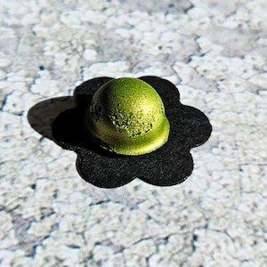 Peut inclure: Un petit objet rond, vert et métallique avec une surface texturée posé sur une découpe en papier noire en forme de fleur.