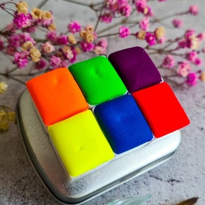 set of six neon palette watercolour paints in a metal tin. The colours are neon: orange, yellow, green, blue, purple, and pink. The tin is sitting on a grey surface with a glass dip pen and a paintbrush.