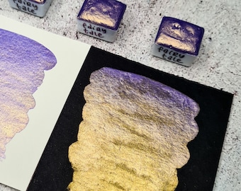 May include: Two swatches of iridescent watercolor paint, one on white paper and one on black paper. The paint is a purple and gold color with a shimmery finish. The paint pans are labeled "Fairy Tale".