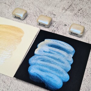 Pode incluir: Uma aproximação de tintas aquarela e amostras. Três pequenas pastilhas de tinta perolada quadradas estão rotuladas como "Funk". Uma amostra de aquarela azul está em papel preto e uma amostra dourada em papel branco.