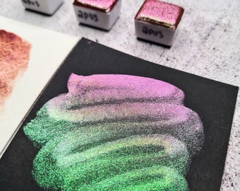 Puede incluir: Tres pequeñas bandejas cuadradas de pintura de acuarela iridiscente de color blanco. Las bandejas están etiquetadas con "APUS". Se muestra una muestra de pintura iridiscente verde y rosa sobre una superficie negra.