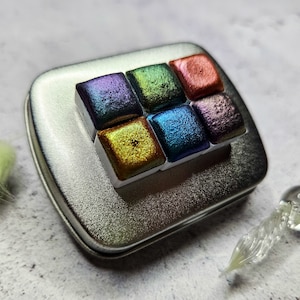 May include: A silver metal tin containing six iridescent watercolour paint pans. The pans are arranged in two rows of three. The colours are purple, green, red, gold, blue, and purple.