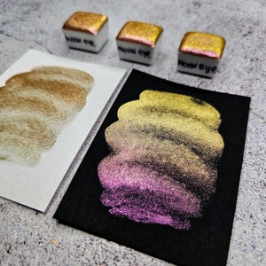Puede incluir: Tres pequeños godets de pintura de acuarela blancos con pigmento metálico dorado y rosa, etiquetados como "moon eye". Se muestran dos muestras de la pintura sobre papel blanco y negro, mostrando un efecto brillante y cambiante de color. La pintura pasa de dorado a rosa.
