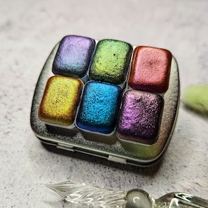 May include: A small, silver metal tin containing six rectangular watercolor paint pans in various iridescent colors: purple, green, red, gold, blue, and violet. A clear glass dip pen and a soft brush are also visible.