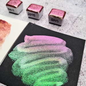 Puede incluir: Tres pequeñas bandejas cuadradas de pintura de acuarela iridiscente de color blanco. Las bandejas están etiquetadas con "APUS". Se muestra una muestra de pintura iridiscente verde y rosa sobre una superficie negra.