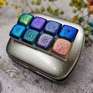 May include: A small silver tin containing a set of watercolour paints. The paints are square and display a spectrum of iridescent colours, including blues, greens, purples, and a shimmering gold. The paints are neatly arranged, indicating a compact art supply.