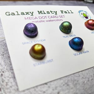 Puede incluir: Un juego de tarjetas titulado "Galaxy Misty Fall" con acuarelas metálicas. La tarjeta muestra cinco muestras de acuarela en forma de puntos en tonos de morado, verde, azul, dorado y rojo. Los colores son "Space System", "Teal Mess", "Solar Rain", "Starburst" y "Meteor".