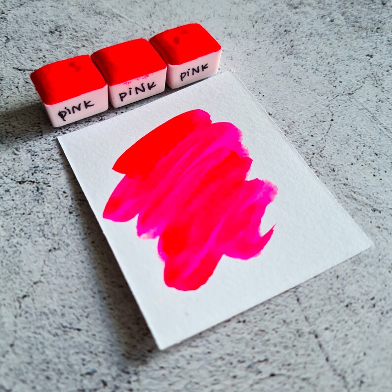 Op de afbeelding: Drie kleine witte blokjes met het woord "PINK" erop geschreven. Een roze verfstaal op een wit vel papier.