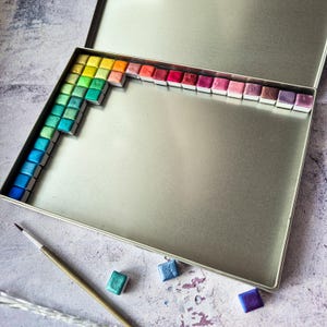 Puede incluir: Una paleta de pintura de acuarela de metal con una variedad de colores dispuestos en un patrón de arcoíris. La paleta está abierta y las pinturas son visibles. También hay algunas pastillas de acuarela individuales sobre una superficie gris.