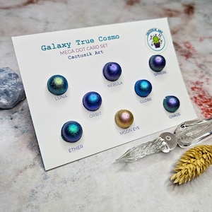 May include: A white card with the text "Galaxy True Cosmo MEGA DOT CARD SET Cactusik Art" and a selection of 10 iridescent paint samples in small round containers. The paint colours are labelled "Luna", "Ether", "Orbit", "Nebula", "Globe", "Sphere", "Moon Eye", and "Chaos".