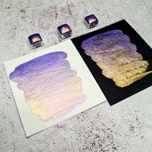 Pode incluir: Close-up de amostras de tinta aquarela em tons de roxo e dourado. Três pequenas pastilhas de tinta quadradas são visíveis, cada uma com texto. As amostras estão em papel branco e preto, mostrando o brilho e a profundidade da cor. O fundo é uma superfície texturizada.