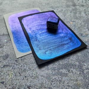 Puede incluir: Dos tarjetas de papel de acuarela con un degradado de tonos morados y azules. Un pequeño cubo oscuro con el texto "Solar Rain" se encuentra encima de una tarjeta. Las tarjetas están sobre una superficie gris texturizada.